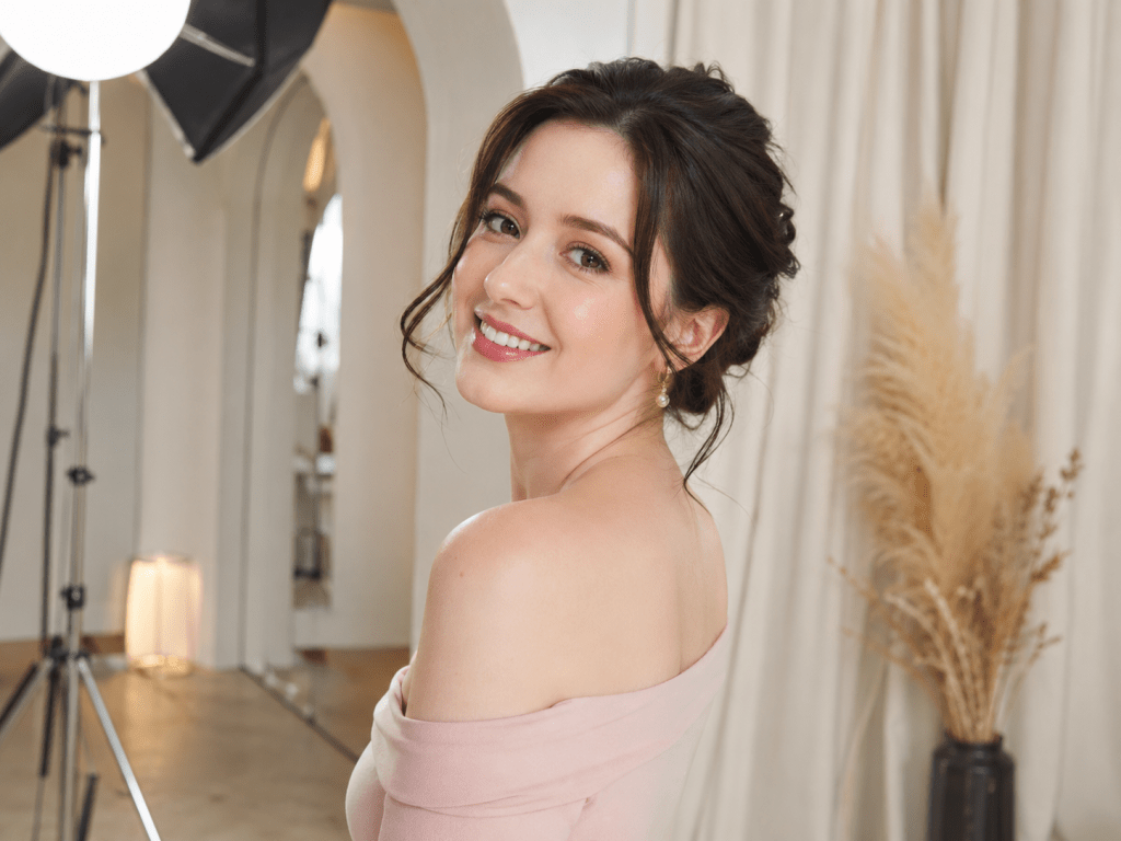 A young woman with long dark hair styled in loose waves smiles at the camera while wearing an off-the-shoulder pink top, in a softly lit studio setting.