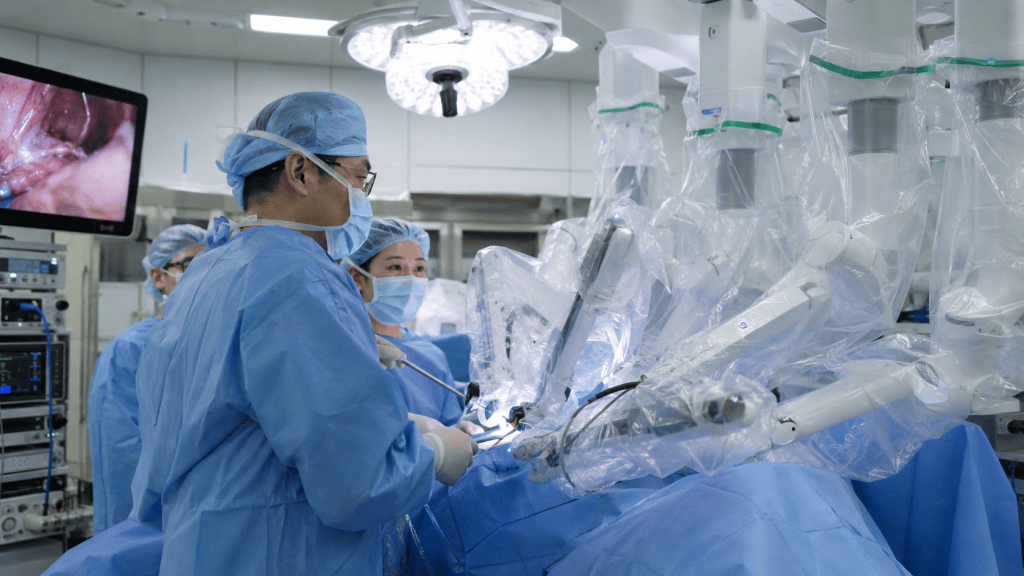Surgeons wearing surgical scrubs and masks performing a procedure with robotic assistance in an operating room, equipped with monitors and surgical instruments.