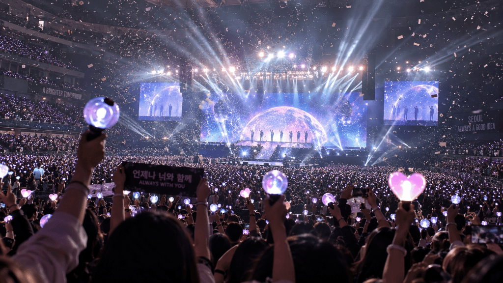 A large crowd at a concert with people holding lightsticks and banners, colorful lights, and confetti in the air, featuring a stage with a cosmic backdrop.