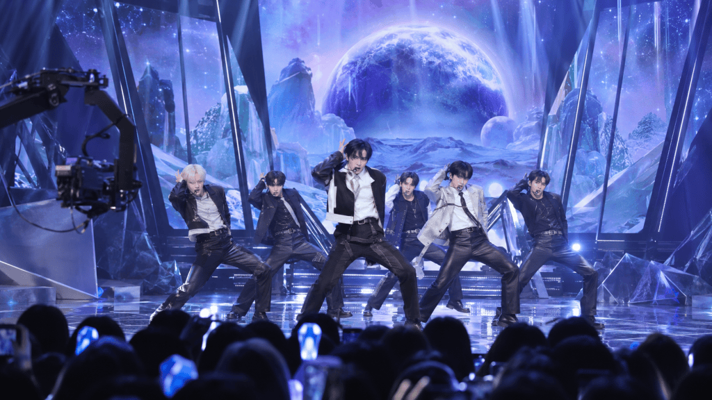 A K-pop group performing on stage with a cosmic backdrop, featuring dramatic lighting and a camera crane capturing the performance.