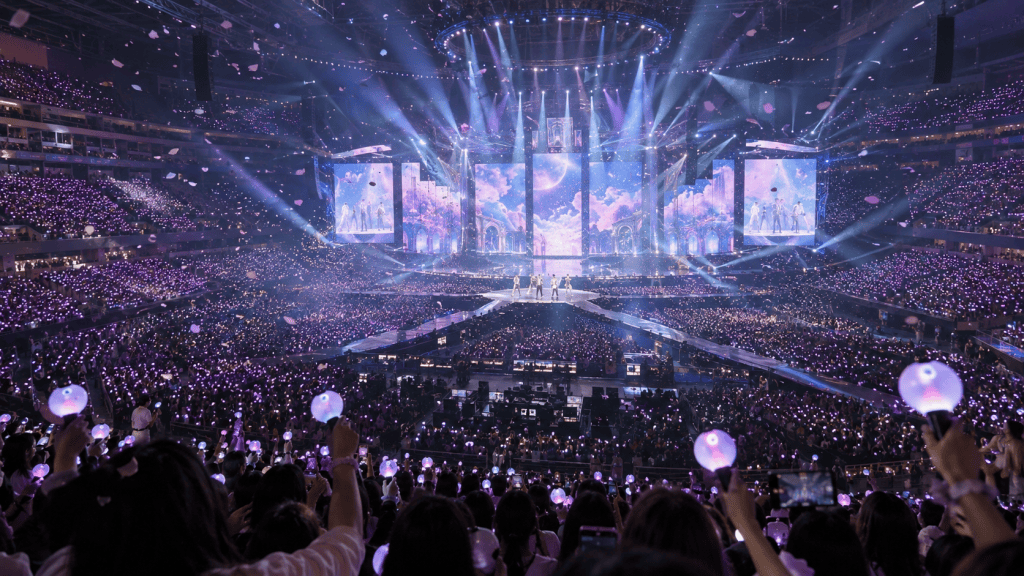 A vibrant concert scene in a large arena filled with fans holding purple light sticks, illuminated by colorful stage lights and a large screen displaying visuals.