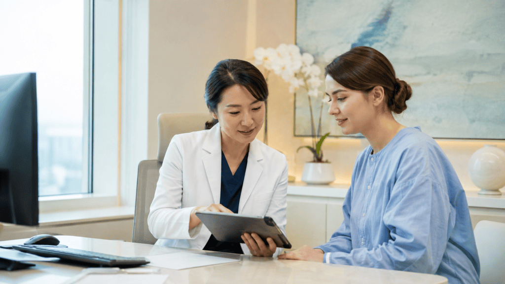 A healthcare professional in a white coat shows a tablet to a patient wearing a blue hospital gown in a modern medical office.