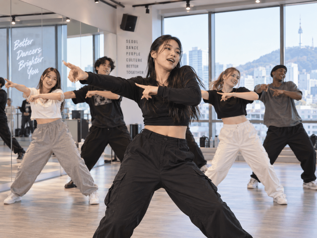 A group of dancers performing in a dance studio with large windows showing a city view. The dancers are wearing casual clothing and are engaged in synchronized movements, showcasing their skills and teamwork.