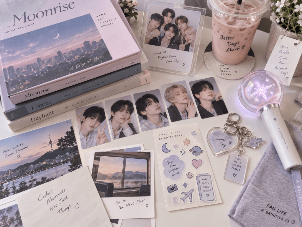 A flat lay composition featuring K-pop inspired merchandise, including photo cards, albums titled 'Moonrise', 'Echoes', and 'Daylight', a pink drink, decorative notes, stickers, and a light stick. The scene has textiles in soft colors and a cozy aesthetic.