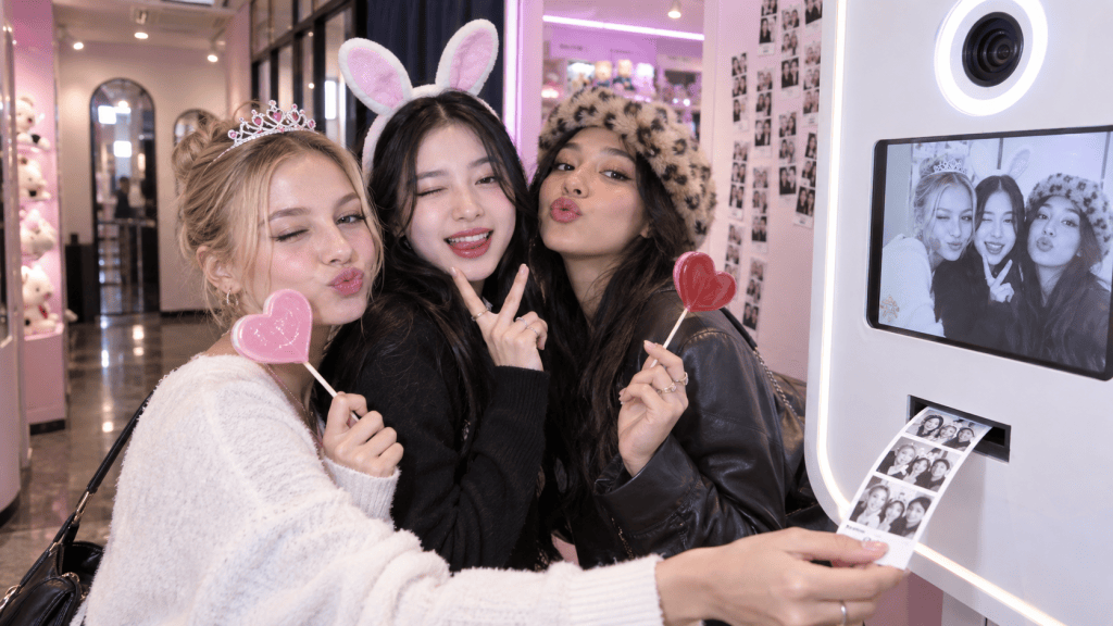 Three friends posing playfully in a fun photo booth with props, including heart-shaped lollipops and bunny ears, while a photo printout displays their cheerful faces.