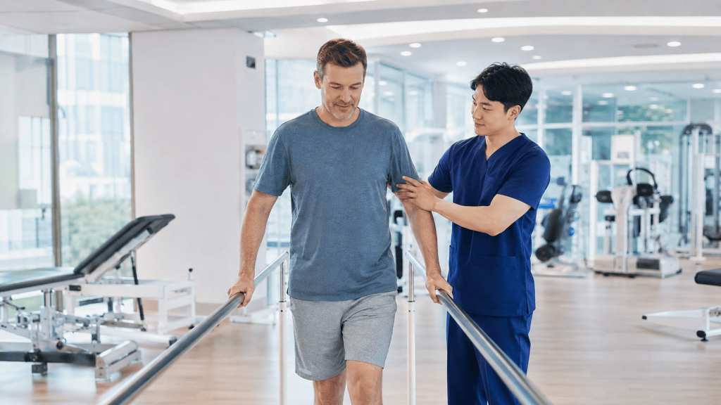 A physical therapist assists a male patient in walking using parallel bars in a modern rehabilitation gym.