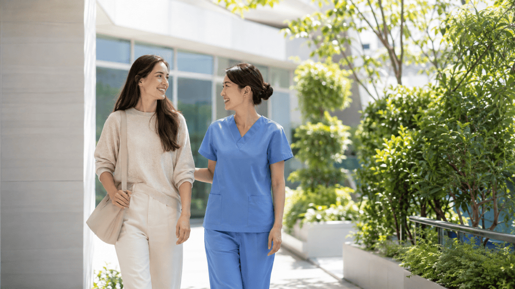 A healthcare professional in blue scrubs and a woman in casual attire walking together outside, surrounded by greenery.