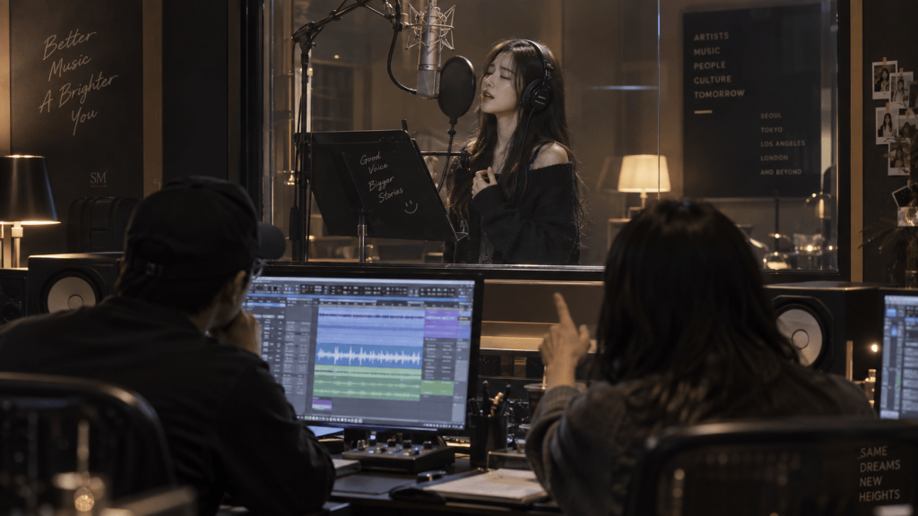 A singer performing in a recording studio, wearing headphones and standing in front of a microphone, while two producers monitor sound on computers.