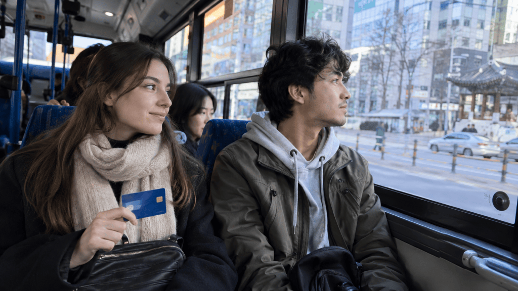 A young woman holding a credit card smiles while sitting on a bus, looking at her companion who gazes out the window. The bus interior shows blue seats and city scenery outside.
