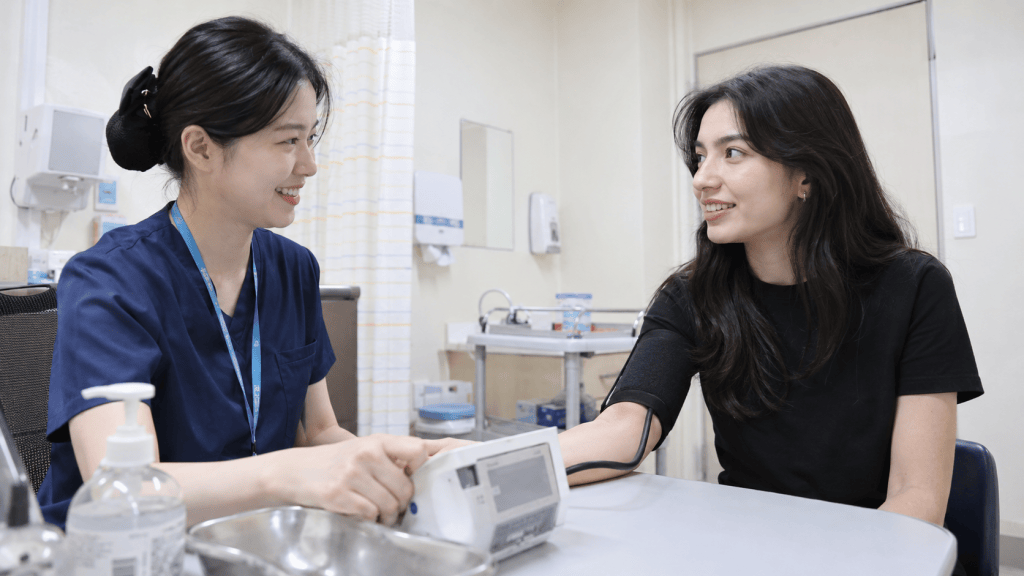 A healthcare professional in blue scrubs takes a blood pressure reading from a young woman sitting at a table in a medical office. Both are smiling and engaged in conversation.