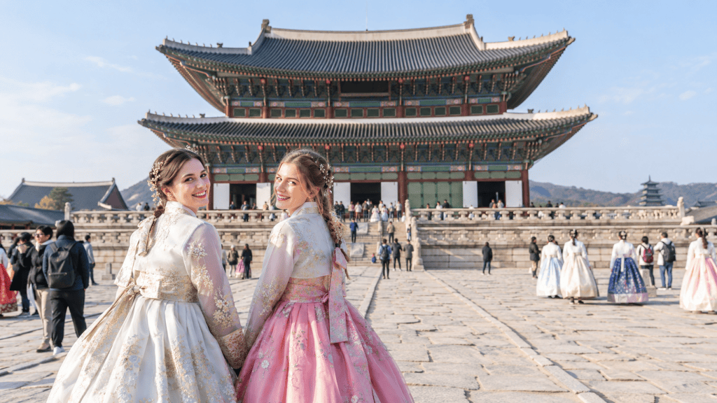 Two women wearing traditional hanbok dresses smile at the camera while standing in front of a historic Korean palace. The background features the palace structure with people walking around.