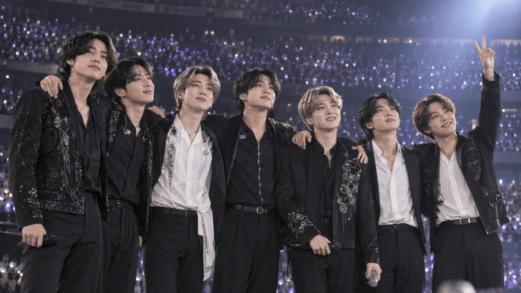 Seven members of a music group standing shoulder to shoulder on stage, smiling and posing with a crowd of fans and purple lights in the background.