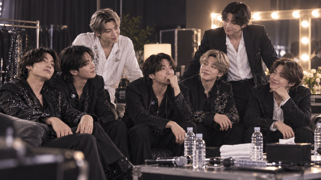 A group of seven men dressed in stylish black outfits, sitting together in a backstage setting, smiling and interacting, with soft lighting and a mirror in the background.