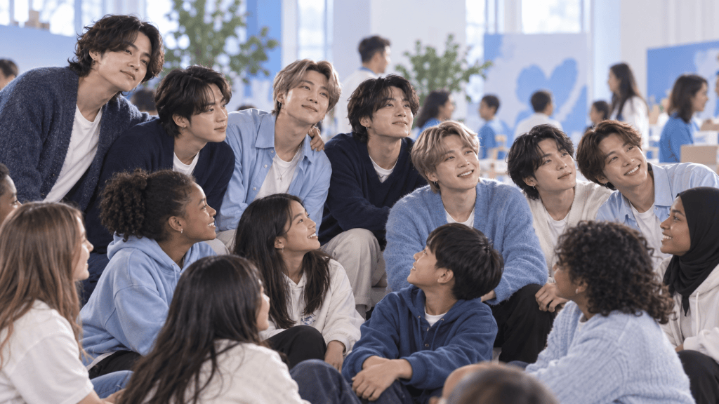 A diverse group of young people, including multiple men and women, sitting together and smiling in a bright, spacious environment with blue and white decor.