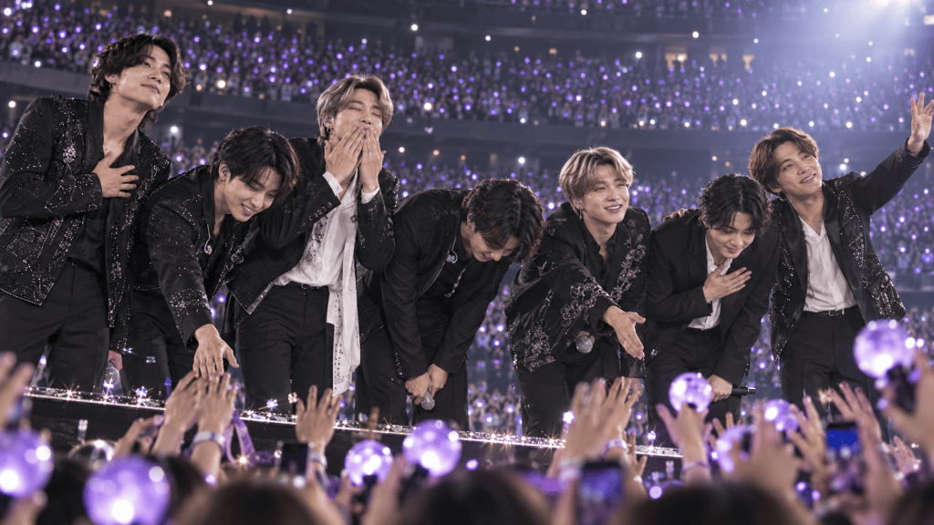 A group of seven male performers on stage, wearing black outfits with sequins, engaging with a crowd holding glowing lightsticks. They appear to be bowing or expressing gratitude, with a vibrant concert atmosphere and a background filled with purple lights.