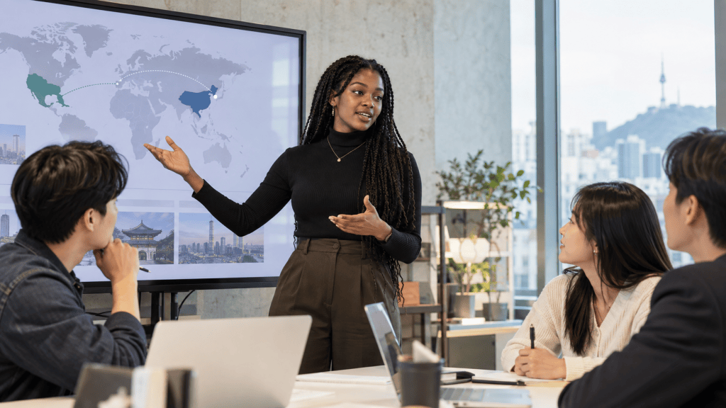 A woman presenting a world map on a screen during a meeting, with three attendees engaged and taking notes.