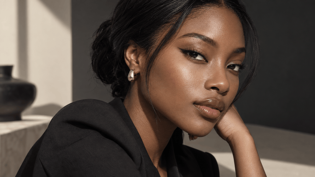 Close-up portrait of a woman with smooth skin and elegant makeup, wearing a black outfit, resting her chin on her hand with a thoughtful expression.