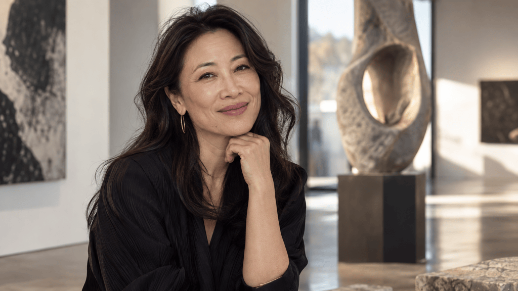 A smiling woman with long, dark hair, wearing a black outfit, poses thoughtfully in an art gallery filled with modern sculptures and abstract paintings.